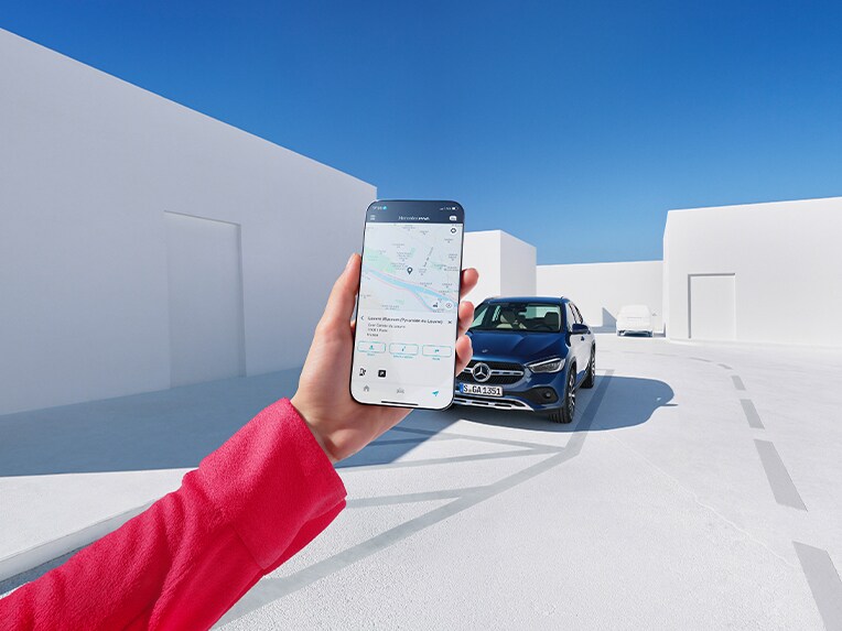 Osoba držící mobilní telefon, na kterém vidíte aplikaci Mercedes-Benz Service. V pozadí stojí vozidlo Mercedes-Benz.