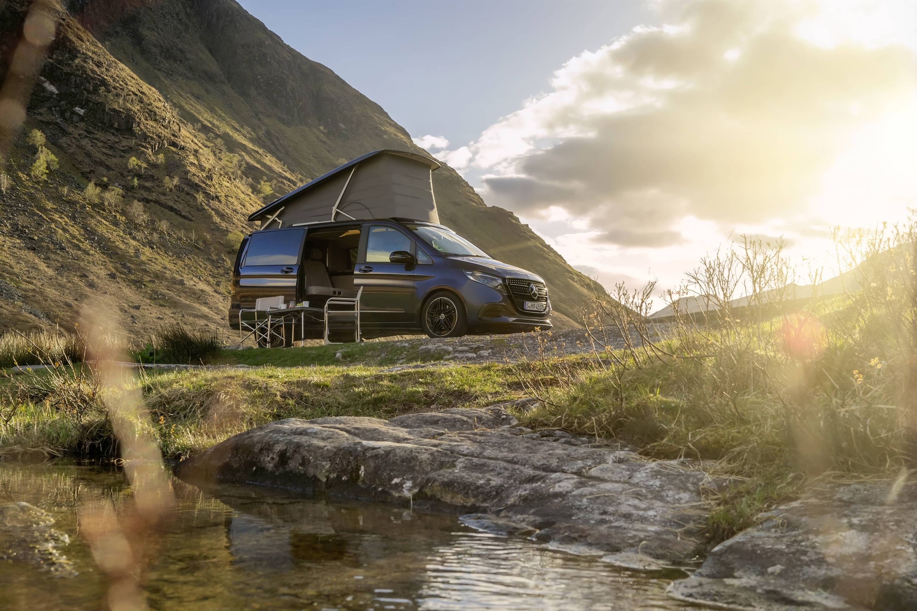 Marco Polo | Cestovní automobily Sprinter | Mercedes-Benz Cestovní automobil Mercedes-Benz Marco Polo s výklopnou střechou zasazený do malebné horské krajiny, demonstrující outdoorové dobrodružství a svobodu života v dodávkovém vozidle.