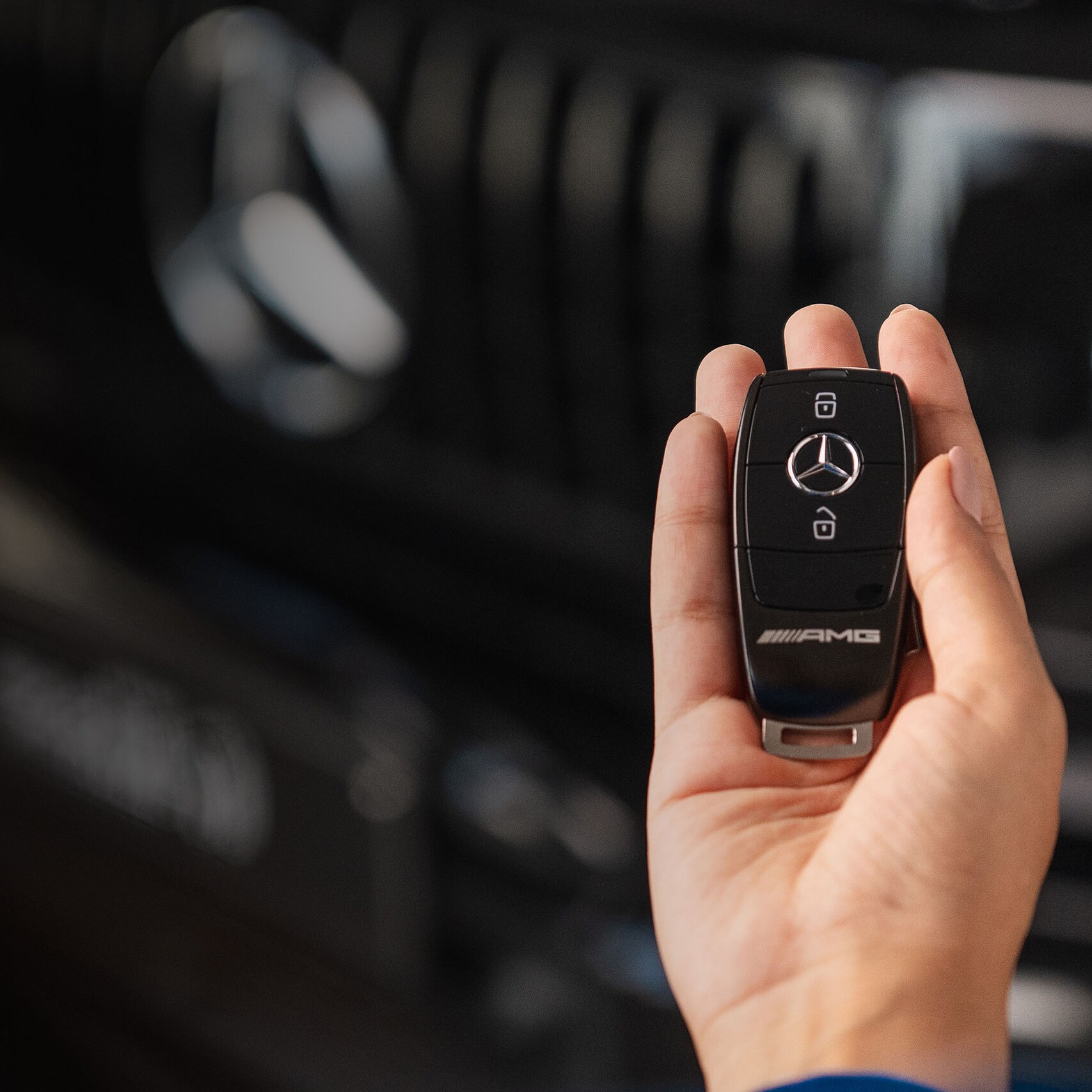 Ruka osoby držící klíčenku k vozidlu Mercedes AMG, s přední maskou vozidla v pozadí.