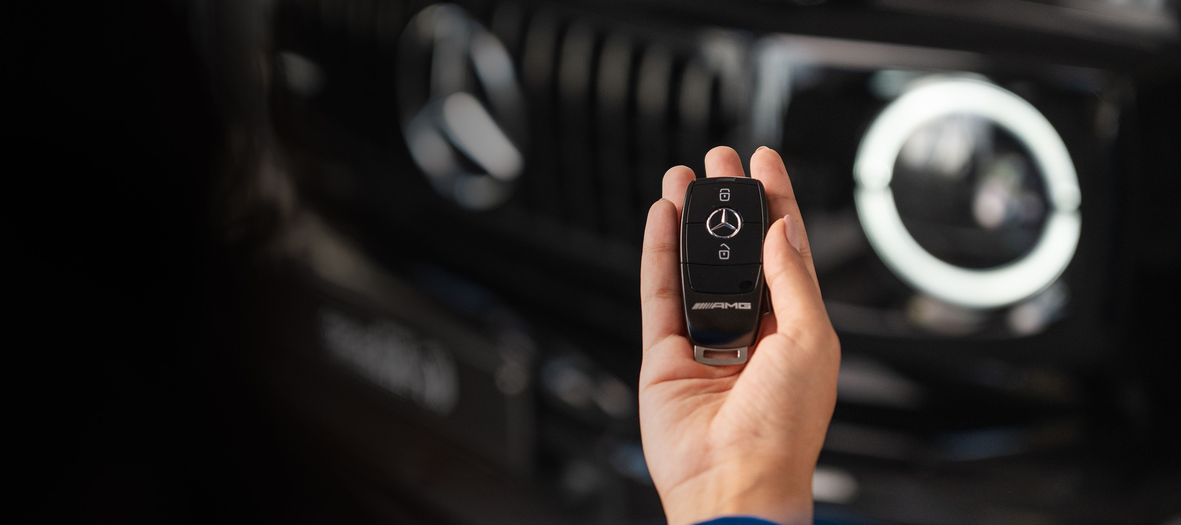 O společnosti Mercedes-Benz Financial Services | Finanční služby | Ruka osoby držící klíčenku k vozidlu Mercedes AMG, s přední maskou vozidla v pozadí.