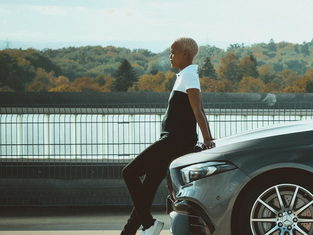 Mladá žena v outfitu z kolekce AMG Racing se nedbale opírá o kapotu Mercedesu.