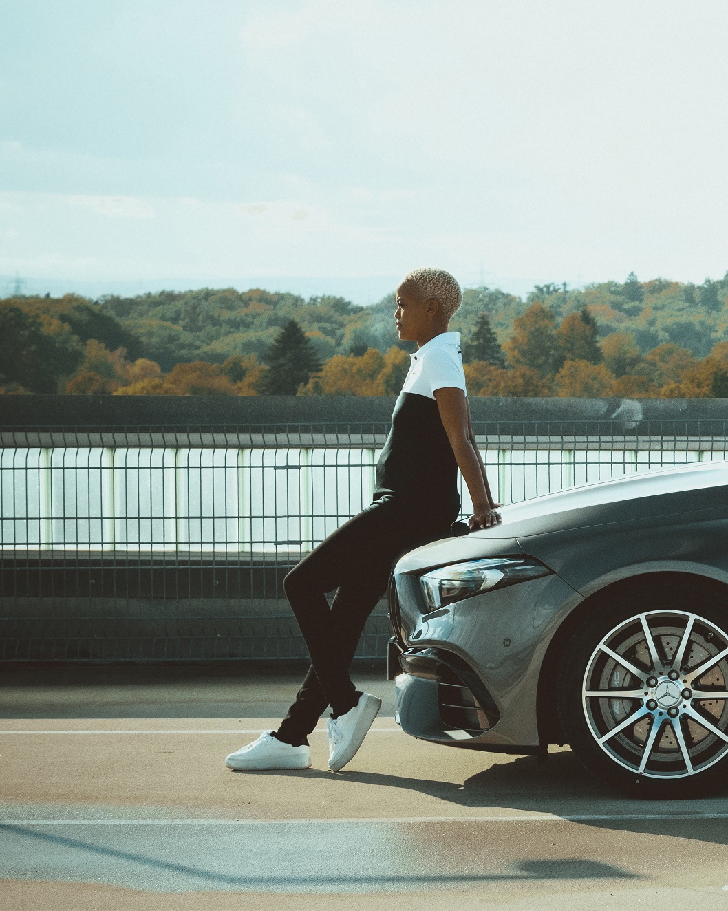 Kolekce AMG Mercedes-Benz | Mercedes-Benz Mladá žena v outfitu z kolekce AMG Racing se nedbale opírá o kapotu Mercedesu.