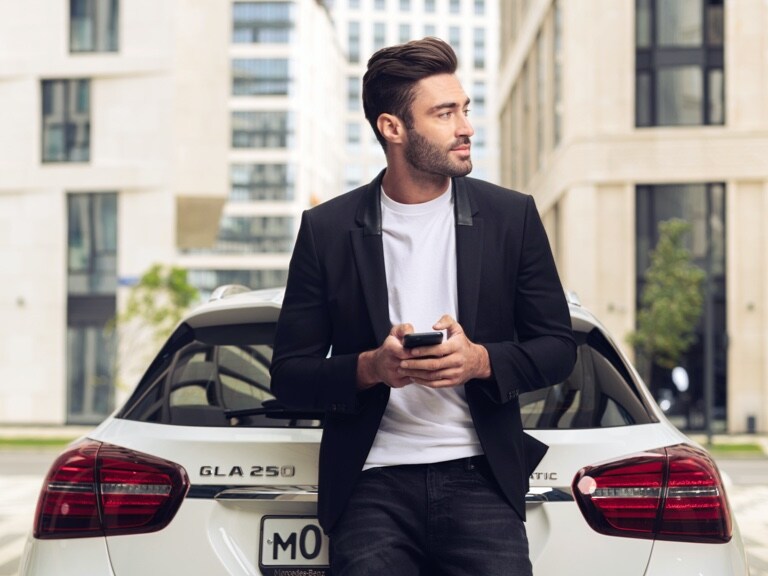 Muž se opírá v sedadle svého vozu Mercedes-Benz GLA a přes aplikaci Mercedes-Benz na svém chytrém telefonu a digitální doplňky k tématu navigace se informuje, jaké bude počasí v místě příjezdu.
