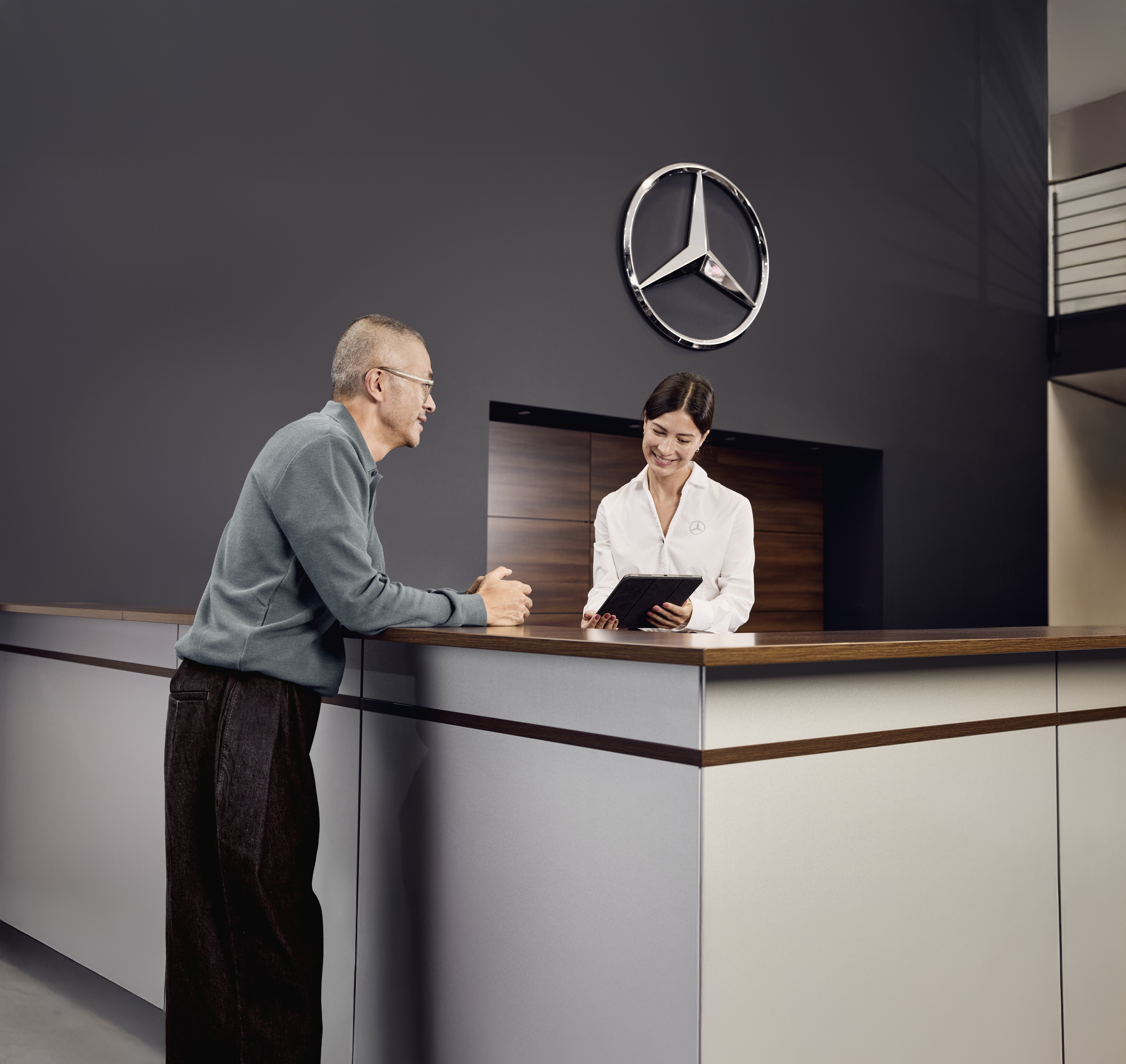 Konzultace ohledně originálních dílů Mercedes-Benz Oblast příjmu vozidel do servisu v autosalonu Mercedes-Benz, kde zákazník probírá podrobnosti týkající se servisu vozidla s přijímacím technikem u stolu.