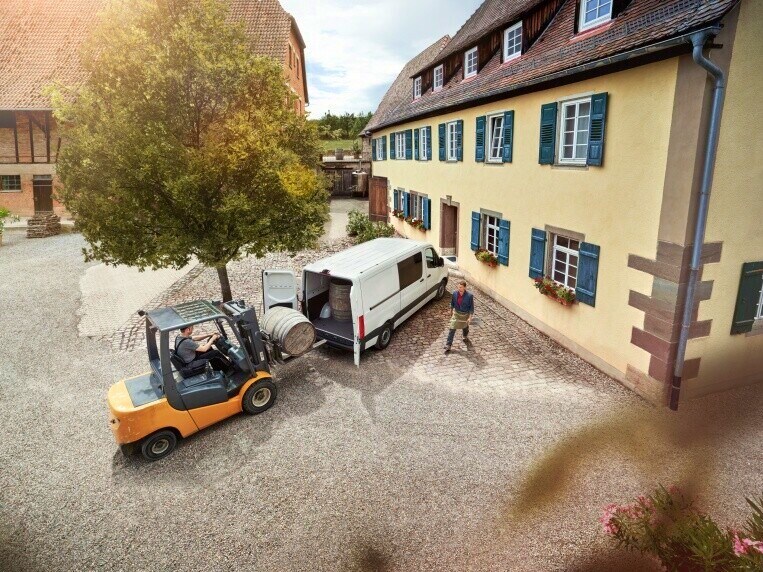 Vysokozdvižný vozík nakládá ve dvoře těžký předmět do skříňové dodávky Mercedes-Benz Sprinter.