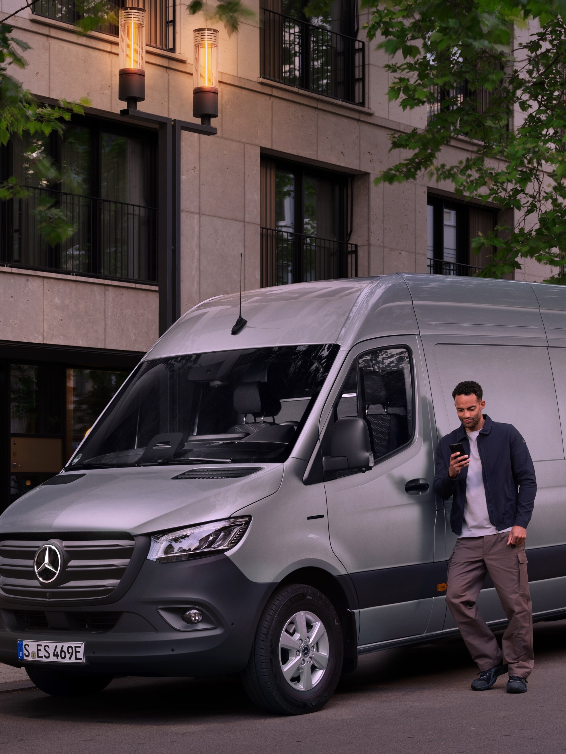 Muž opírající se o stříbrné dodávkové vozidlo Mercedes-Benz Sprinter a používající svůj chytrý telefon – rychlá asistence od zákaznické péče Mercedes-Benz.