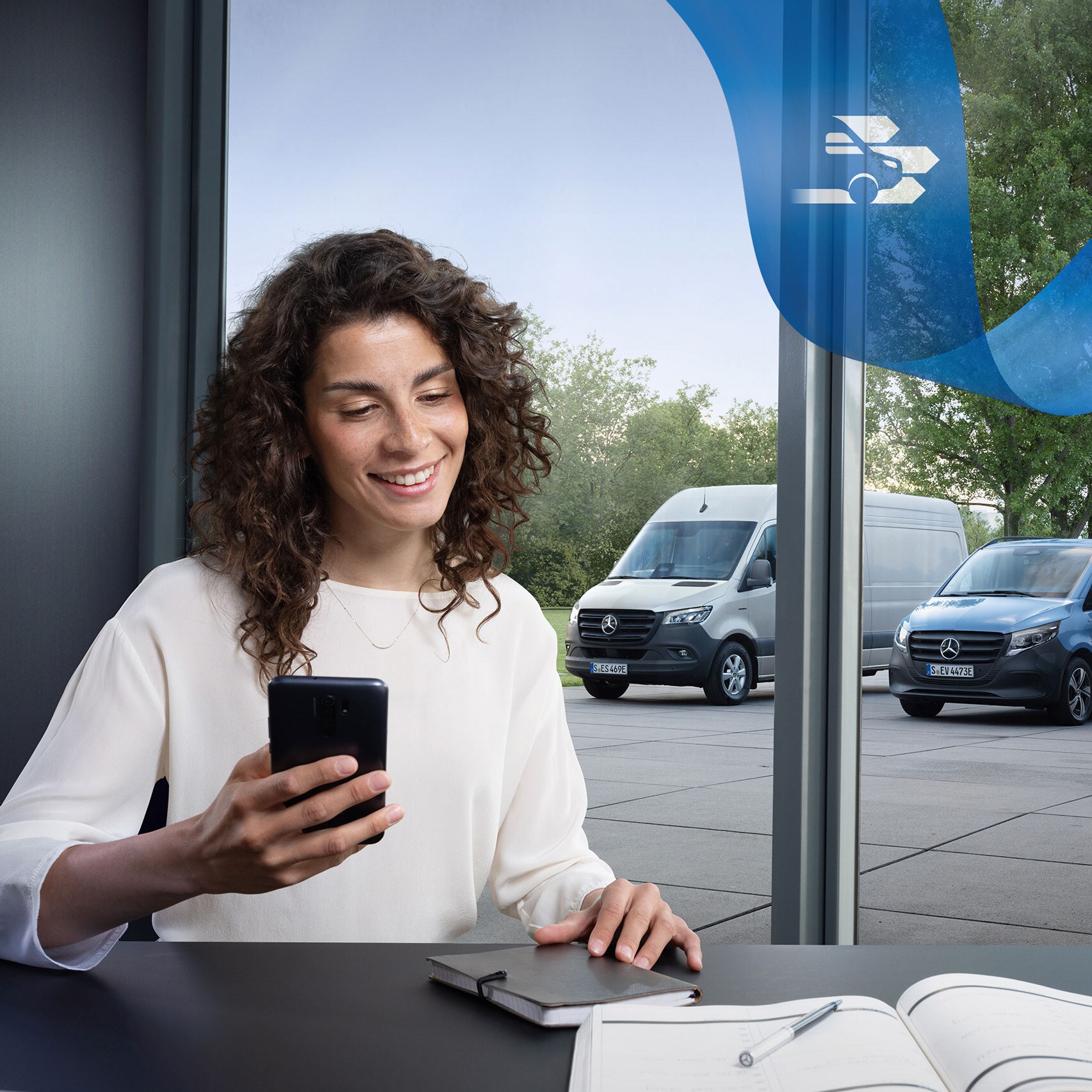 Žena sedící u&nbsp;stolu s&nbsp;chytrým telefonem a&nbsp;v&nbsp;pozadí řada dodávkových vozidel Mercedes-Benz.