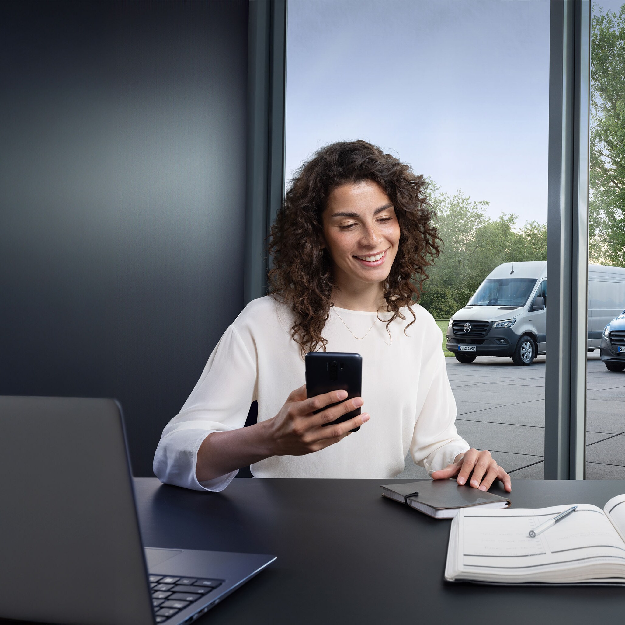 Woman looking for a Mercedes-Benz Service Partner on her smartphone with a range of Mercedes-Benz vans in the background.