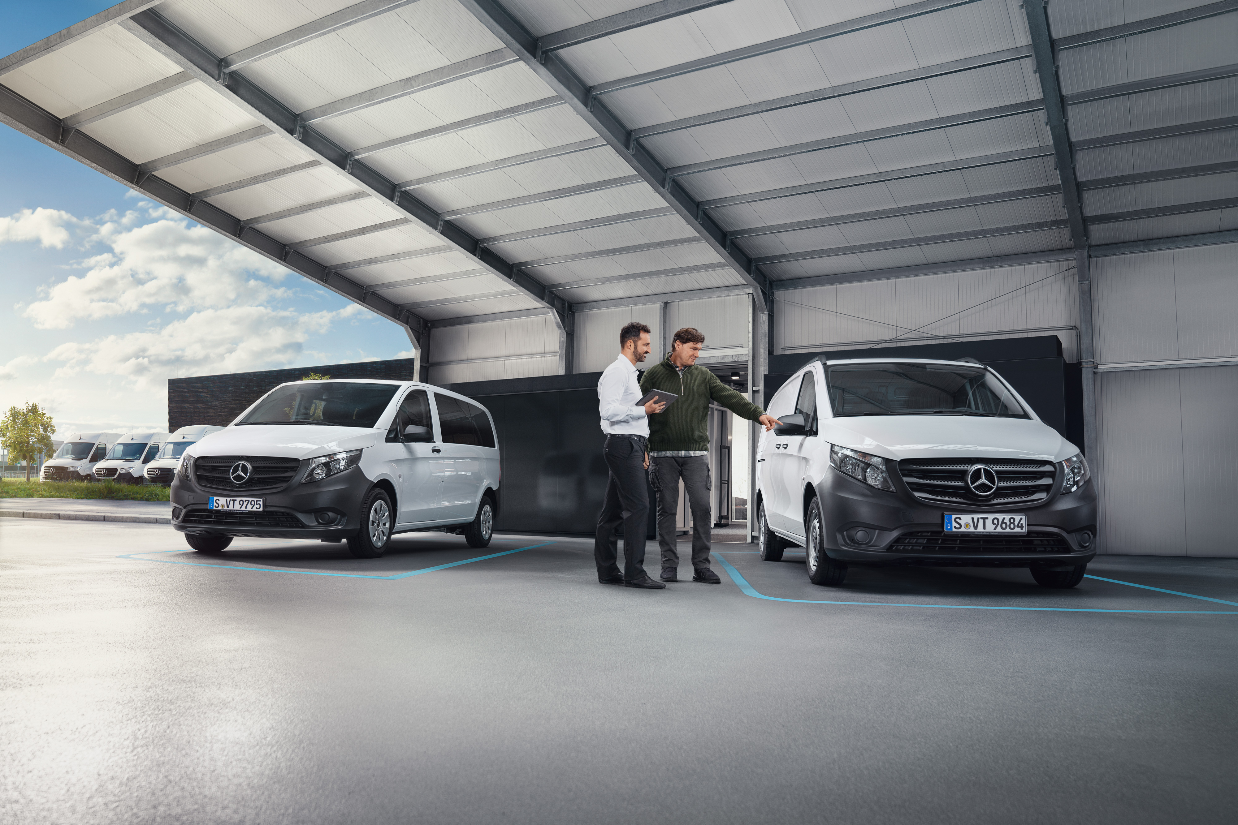 Mercedes-Benz | Pohodlí | Digitální příjem pro zákazníky Dva lidé si povídají v dílně, přičemž stojí před dvěma bílými dodávkovými vozidly Mercedes-Benz.