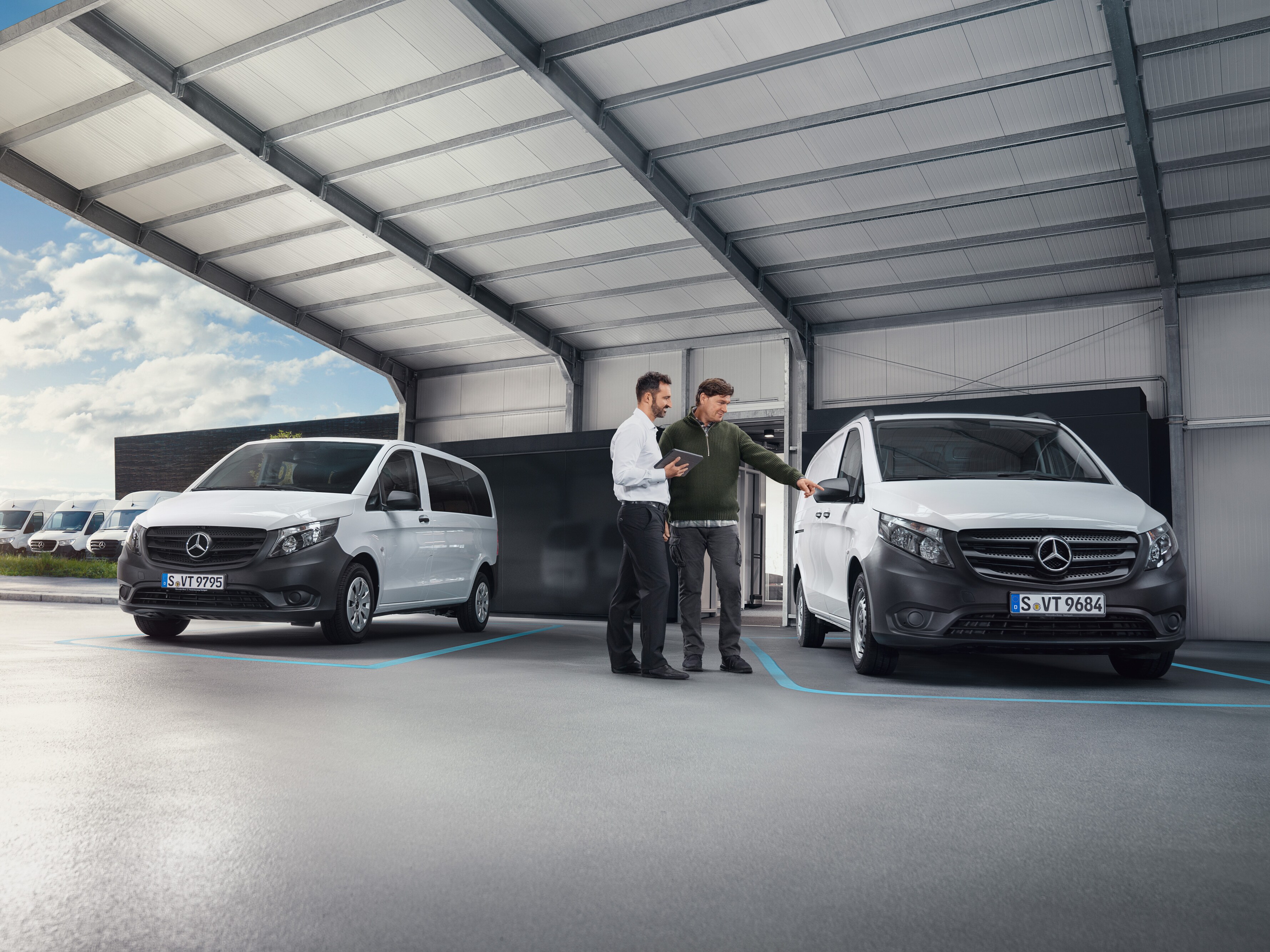 Dva lidé si povídají v&nbsp;dílně, přičemž stojí před dvěma bílými dodávkovými vozidly Mercedes-Benz.
