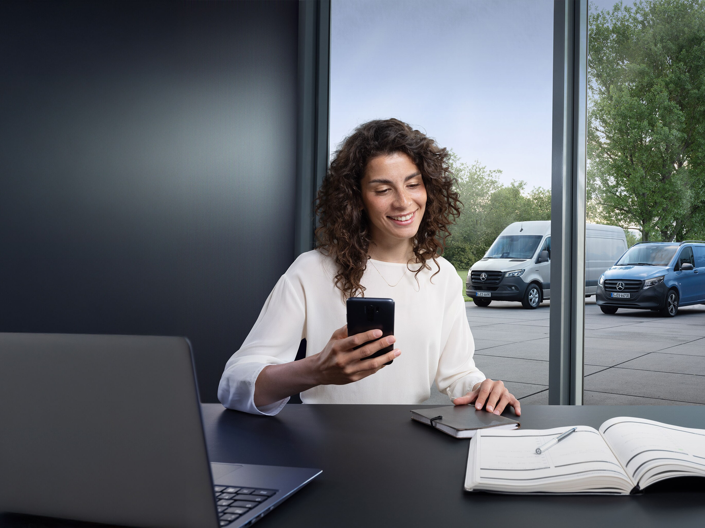 Žena si rezervuje servis přes chytrý telefon s&nbsp;řadou dodávkových vozidel Mercedes-Benz v&nbsp;pozadí.