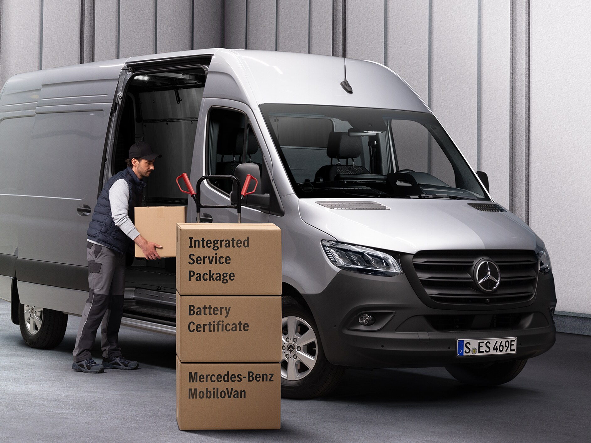 Osoba vykládající krabice z vozidla Mercedes-Benz eSprinter. Před dodávkovým vozidlem jsou naskládány tři krabice. Krabice jsou označeny názvy servisních produktů Mercedes-Benz.