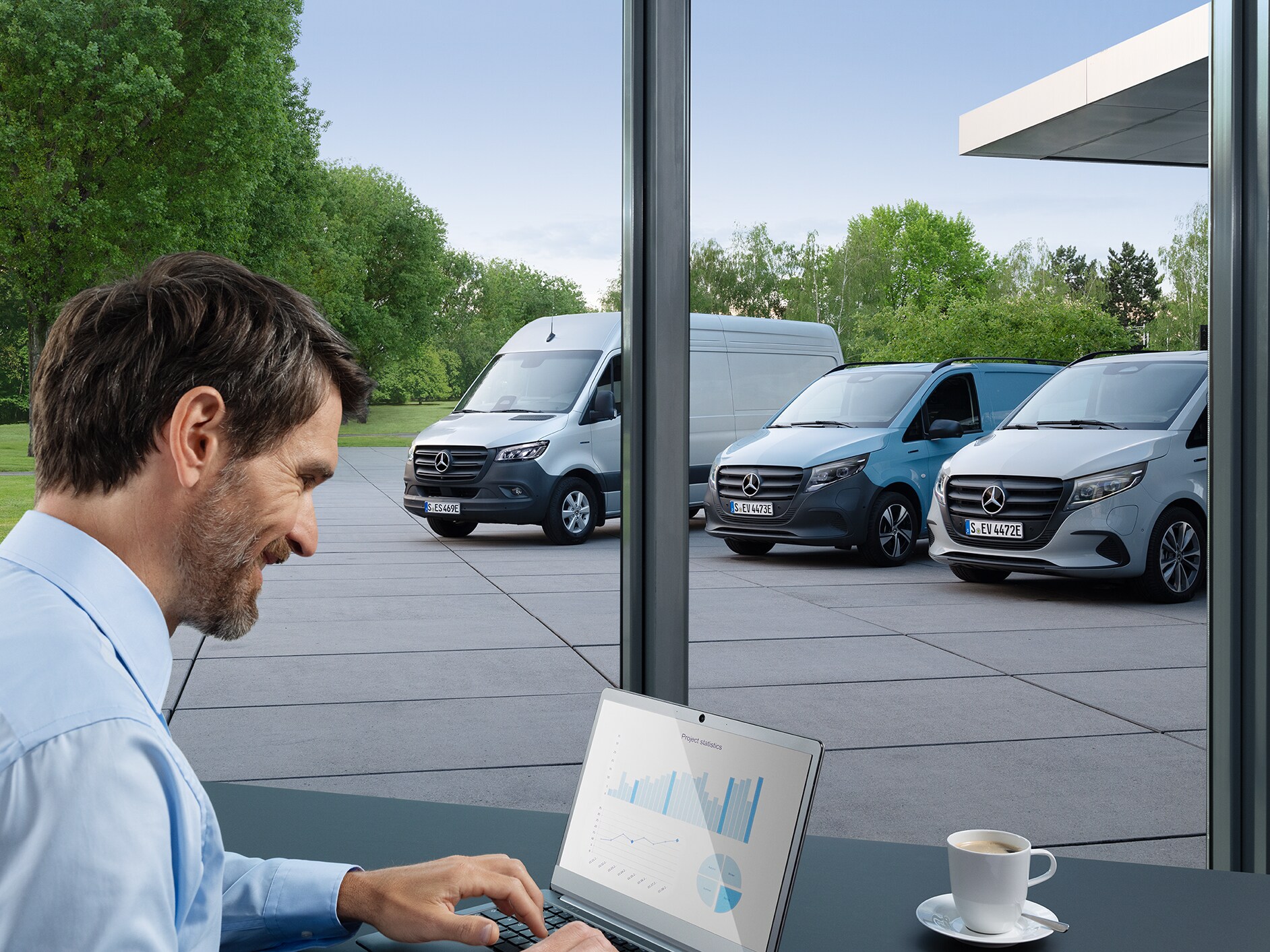 Osoba sedící před laptopem v kanceláři. Venku jsou zaparkovaná tři elektrická dodávková vozidla Mercedes-Benz.
