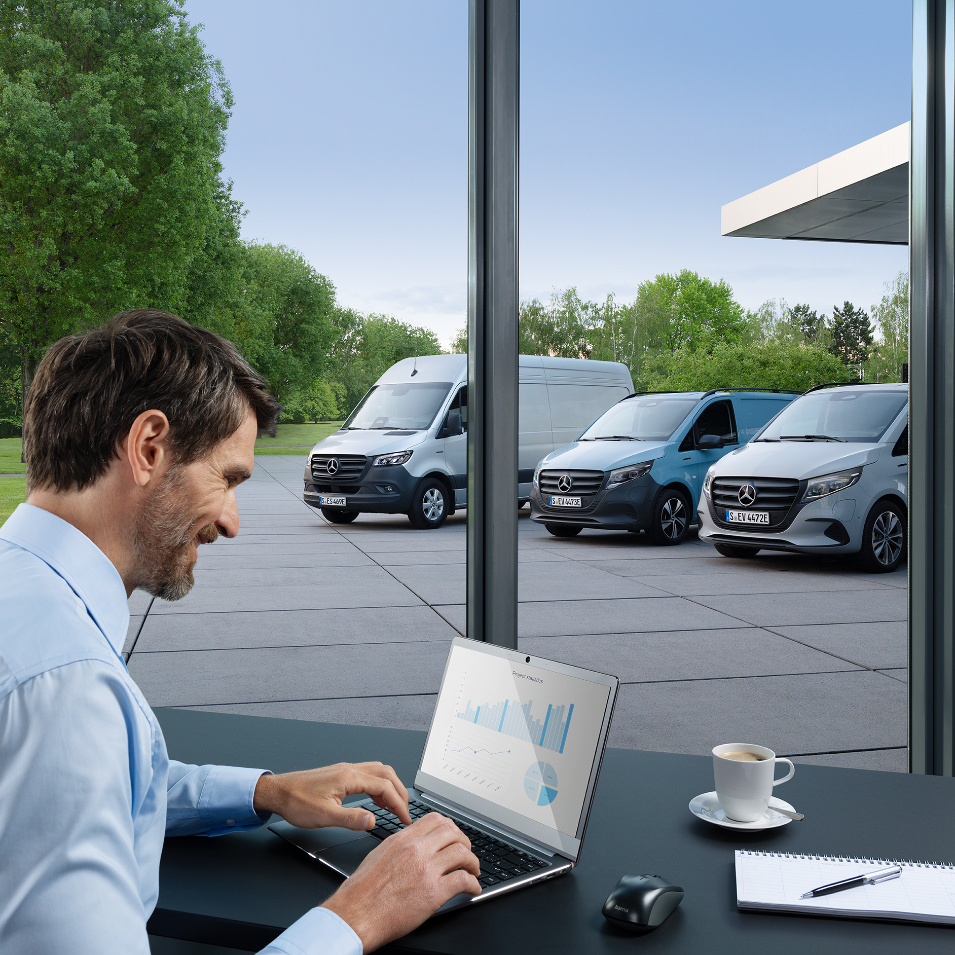 Elektromobily Mercedes-Benz | Mercedes-Benz ServiceCare Osoba sedící před laptopem v kanceláři. Venku jsou zaparkovaná tři elektrická dodávková vozidla Mercedes-Benz.