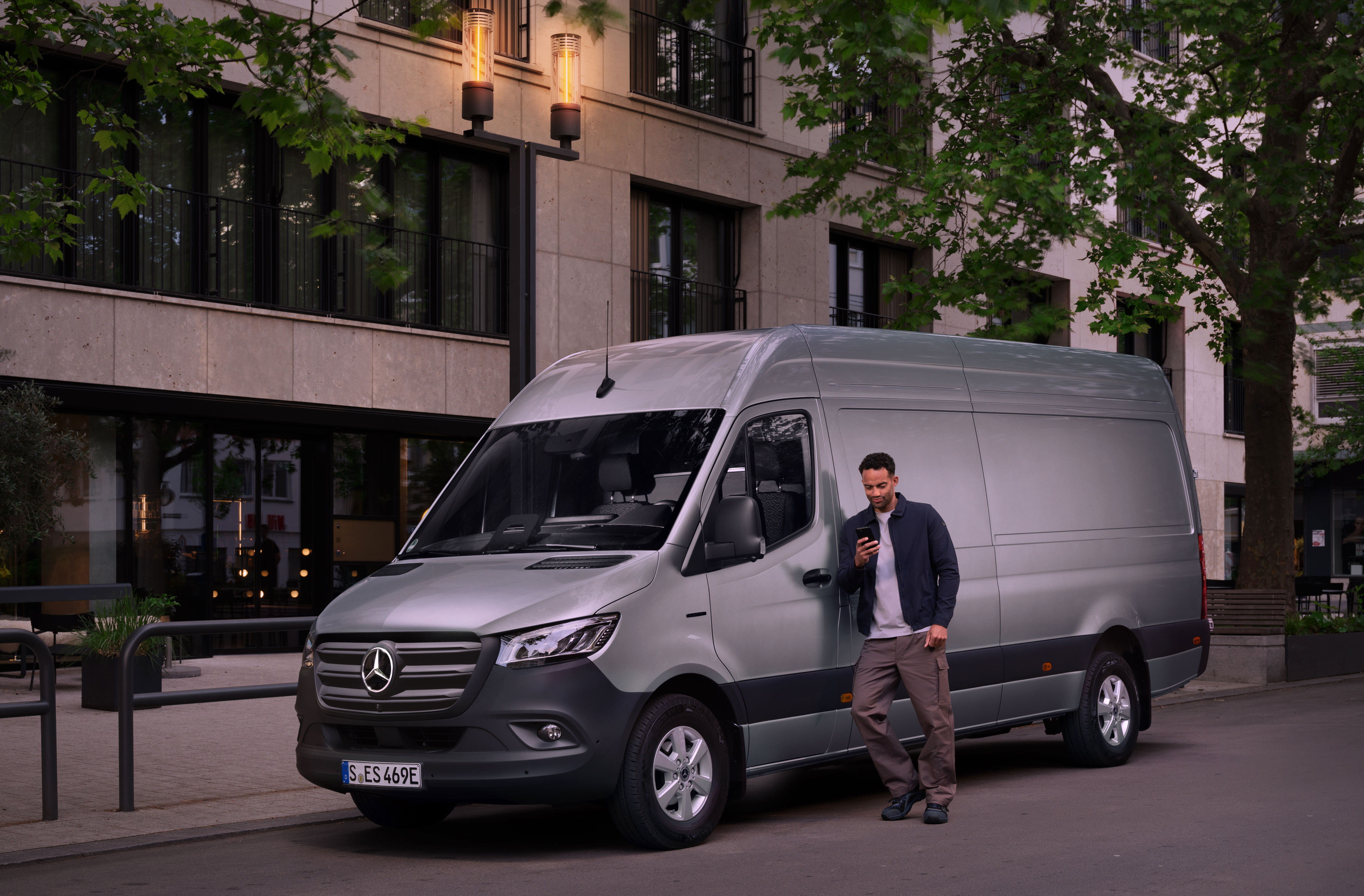Mercedes-Benz | Oprava | Digitální zpráva o škodách Muž se opírá o stříbrný Mercedes-Benz Sprinter a vyplňuje zprávu o škodách na svém mobilním telefonu.