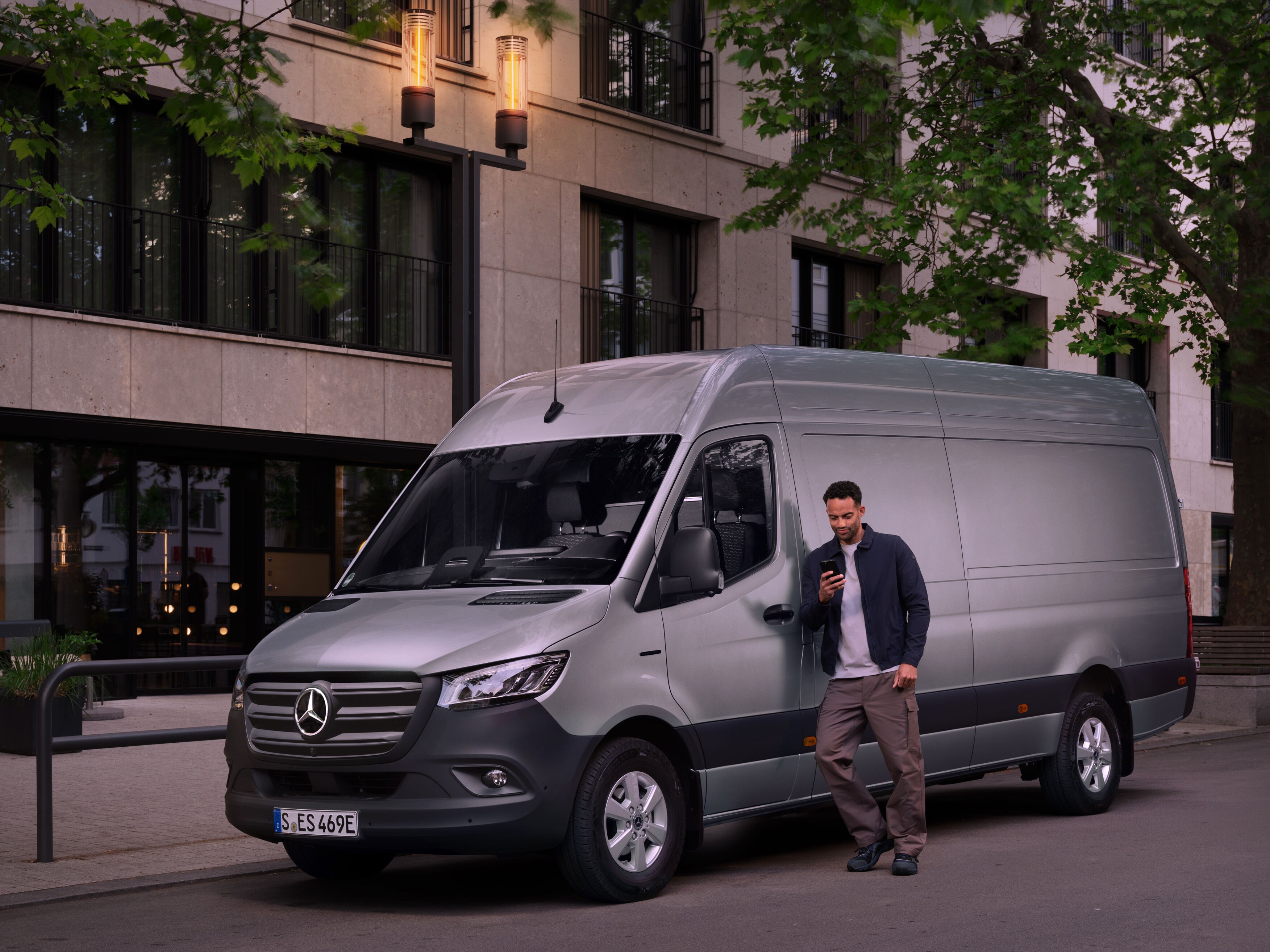 Muž se opírá o&nbsp;stříbrný Mercedes-Benz Sprinter a&nbsp;vyplňuje zprávu o&nbsp;škodách na svém mobilním telefonu.