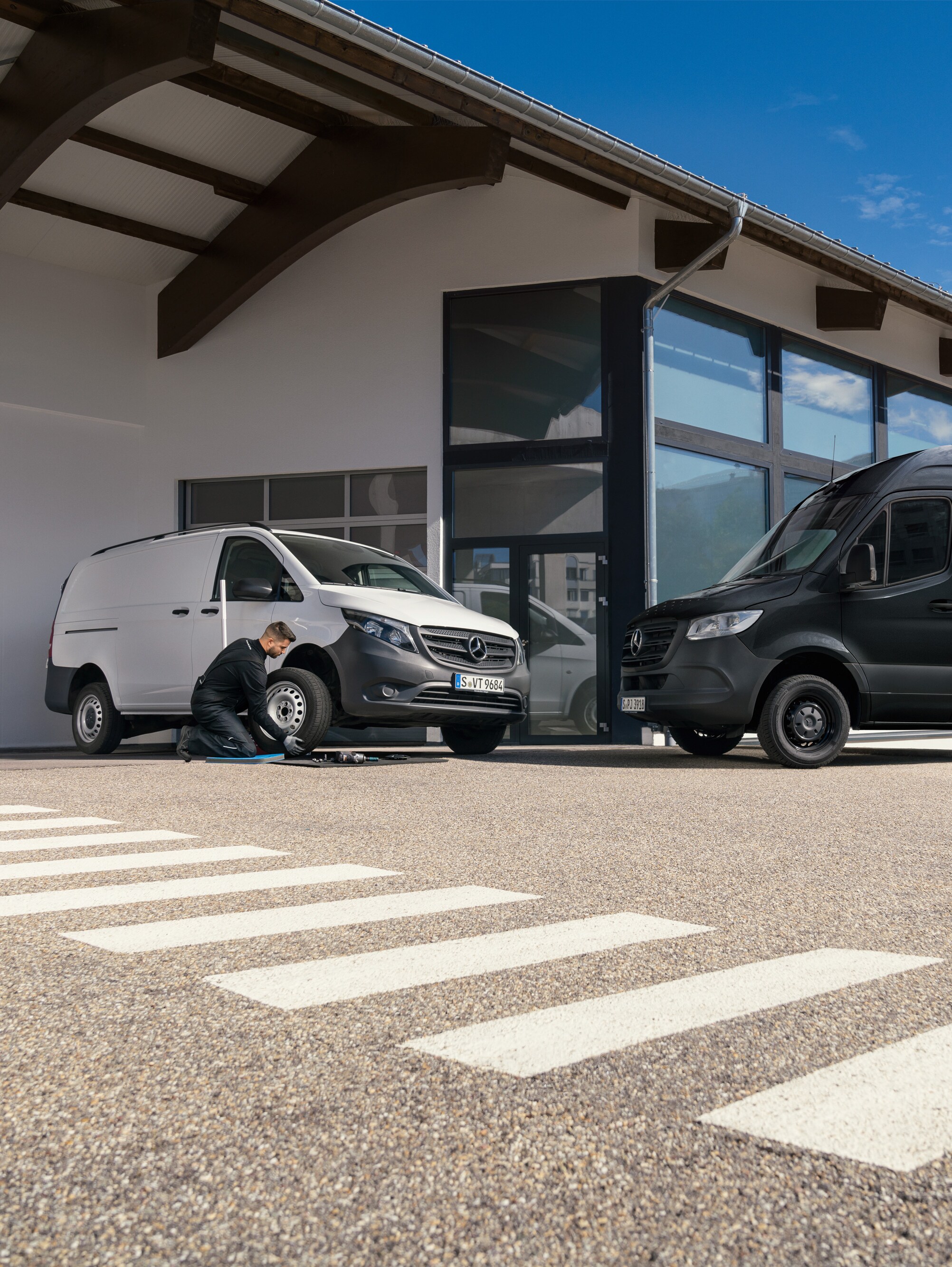 Technik mění pneumatiku na bílém dodávkovém vozidle Mercedes-Benz Vito před showroomem.