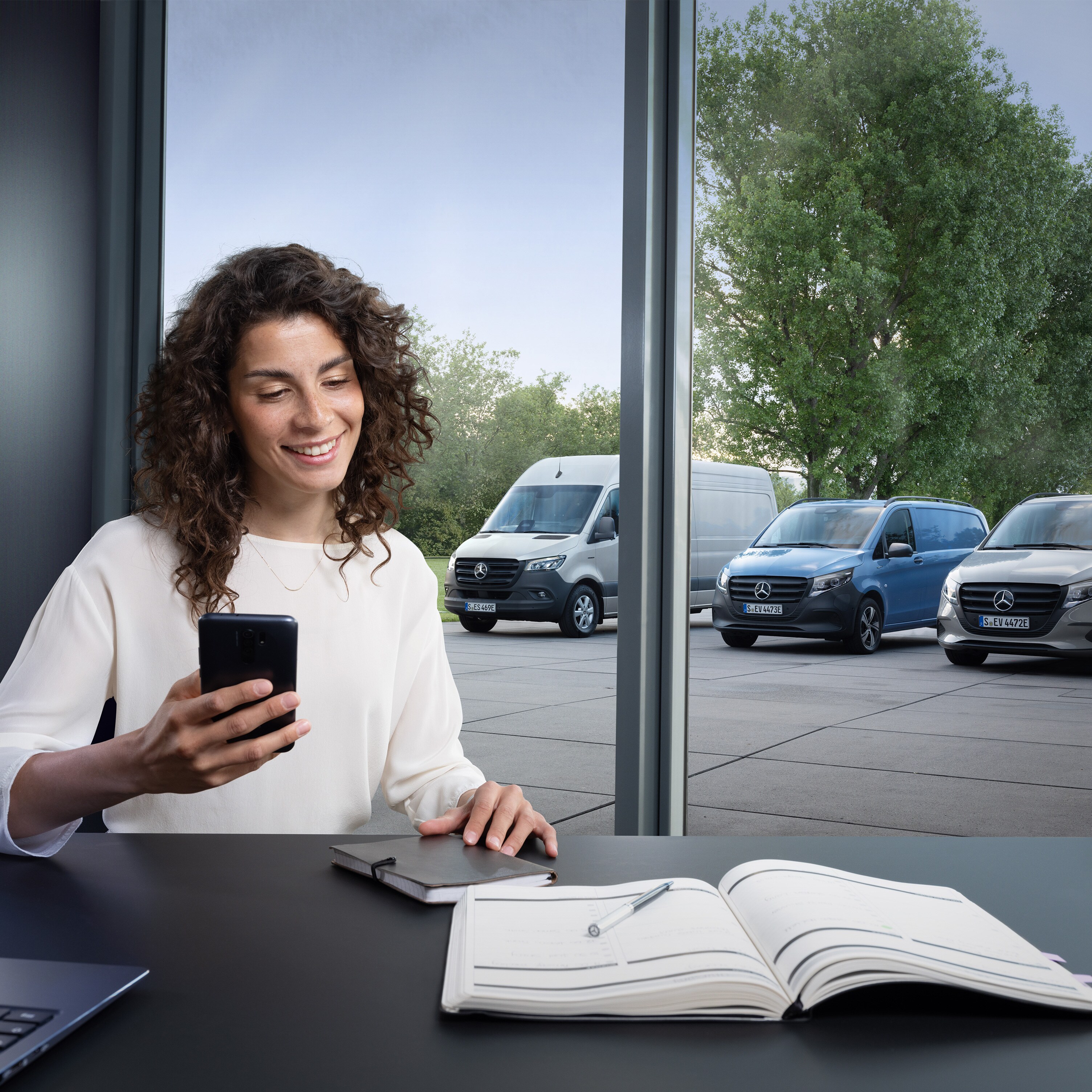 Žena dělá plány na svém chytrém telefonu před řadou dodávkových vozidel Mercedes-Benz v&nbsp;pozadí.